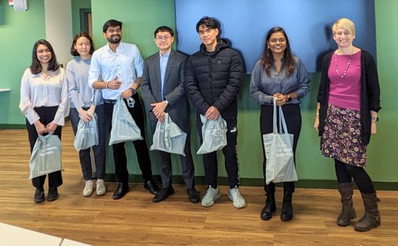 A diverse team of seven MBA students holding grey tote bags standing in front of a large TV mounted on a green wall.	