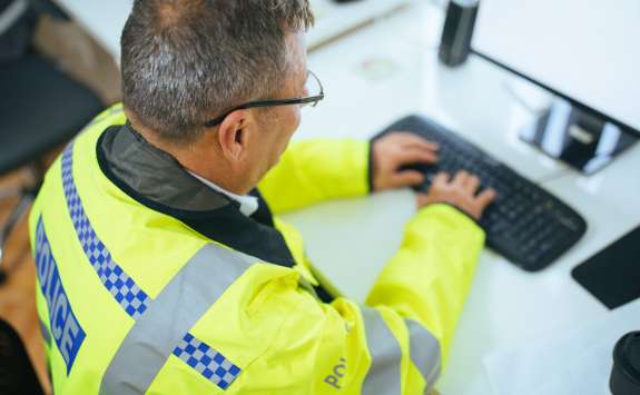 A policeman in a yellow hi-vis police jacket typing at a computer keyboard. 