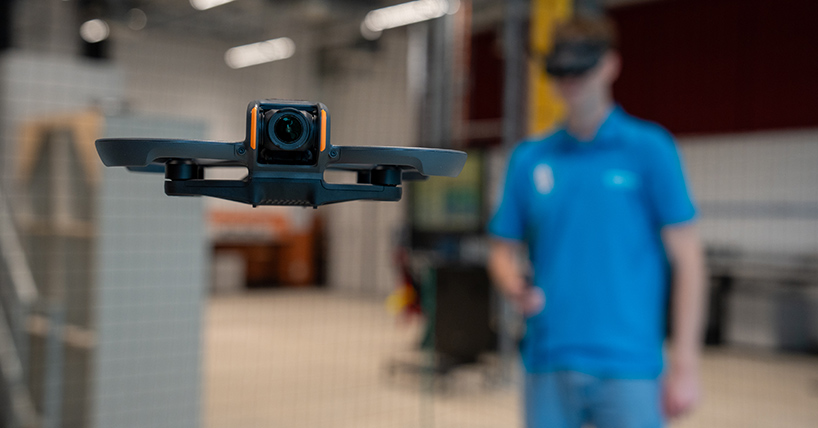 A student in a blue t shirt with a headset on guiding a drone flying in the foreground