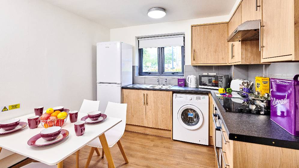 A small kitchen that has a square table which is set for four people. There is a window above the sink. There is also an electric hob, a washing machine, kettle, toaster and microwave.
