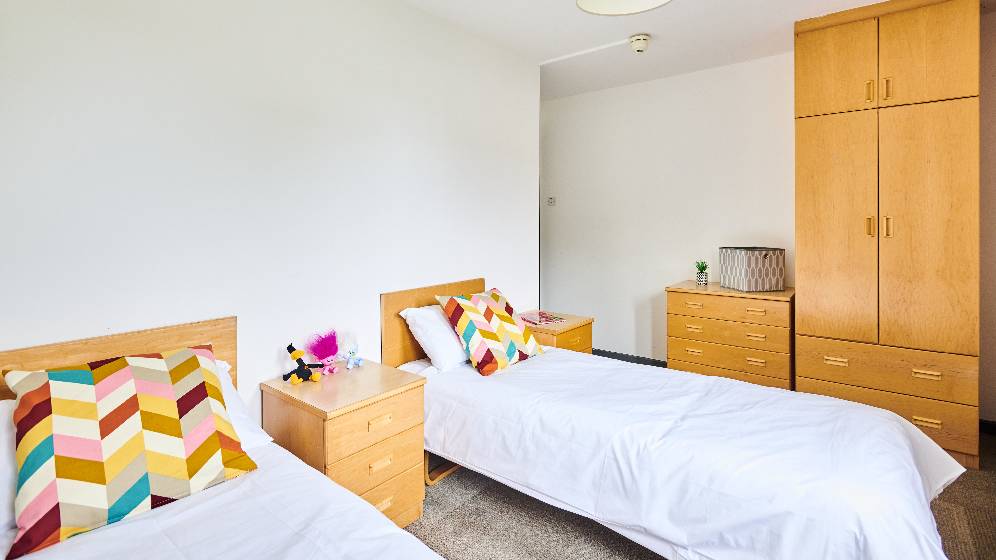A bedroom with two single beds. There is a bedside table between the two beds which have children's toys on. There is an additional bedside table, drawers and a wardrobe.