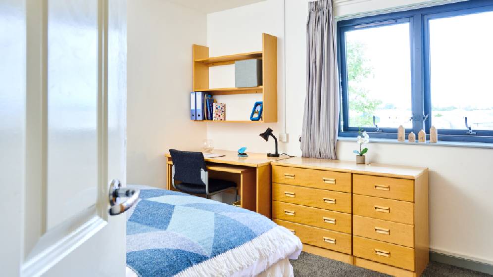 A photo taken from the doorway of a bedroom. It shows the corner of the bed, a desk, a chair, shelves, drawers and a window.