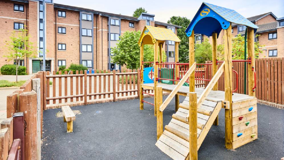 A small wooden children's adventure playground. There is a slide and various other ways to climb onto the structure. There is a fence surrounding the park, and a bench to sit on.