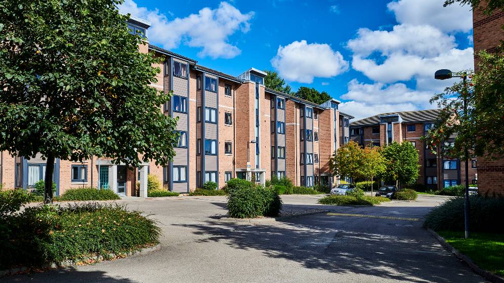 The exterior of Bowsden Court. The building is four stories high. It is a sunny day and there are green trees and bushes in front of the building