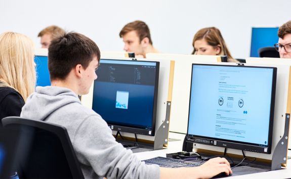 Students are working at computer work stations. The workstations are "flip top" desks. The desks can be opened to reveal a PC and Raspberry Pi machine. Or the desks can be flipped to a plan desktop so that students can use their own laptops and tablets.