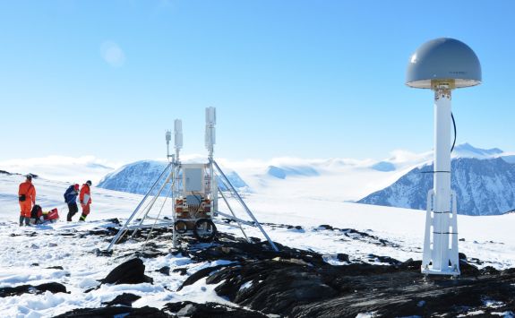 Researchers are working in an icefield. There is a rocky outcrop. Attached to the rock is research equipment. The main piece of equipment is a metal pole with a dome on top. The dome has the circumference of a large dinner plate. Inside the dome there is a GNSS receiver antenna.