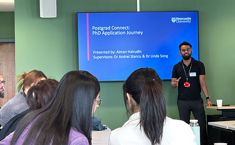 Aiman Hairudin presenting to a group of students at Newcastle University. The screen behind him reads "Postgrad Connect: PhD Application Journey," with his name and the names of his supervisors.