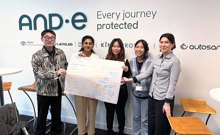 Five students stand in front of an AND-E company wall display, smiling and holding a handwritten presentation poster with key points and colourful annotations.