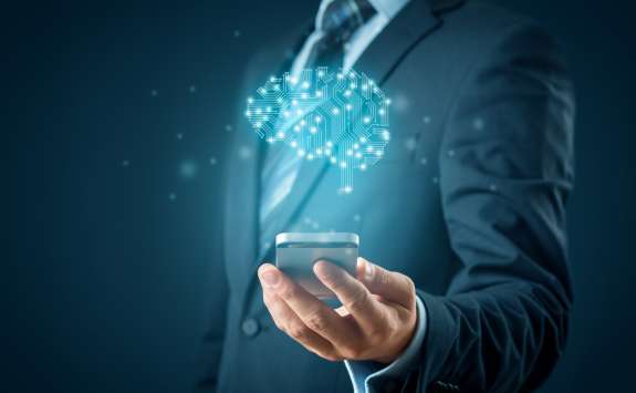 A man in business dress holding a smartphone out in front of him. A blue holographic image of a brain made up of circuit-style lines hovers above it.