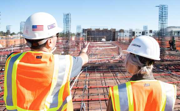 A team leader and colleague in orange high vis vests and white hardhats overlooking a large building site.