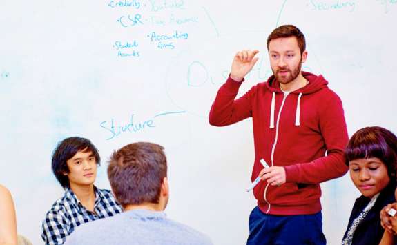 A Business School student in a red hoodie standing in front of a whiteboard, in discussion with other students sat around a table