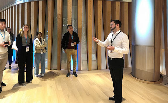 Cameron Bateman leading a tour at Bloomberg, speaking to a diverse group of students in a modern office environment with wooden panel walls and bright lighting. Students listen attentively while standing in a semi-circle around the speaker.	