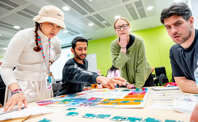 Four students gathered around a board game in a classroom, actively discussing and pointing to different game pieces.