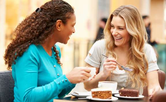 Two women enjoying coffee and cake at a cafe, smiling and engaged in a lively conversation, one woman is sharing her cake with the other.