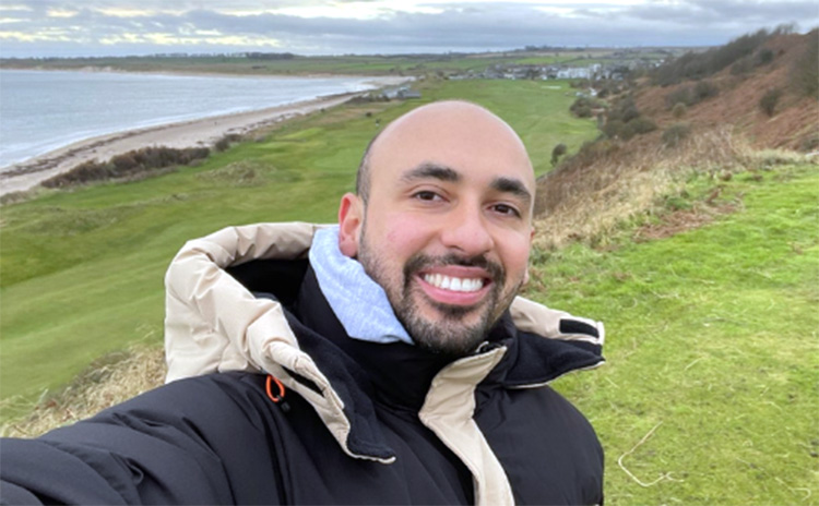Erminsson Tirado Delgado wearing a black and beige winter jacket smiles at the camera while taking a selfie on a grassy coastal hill. The sea, beach, and countryside stretch into the distance under a cloudy sky.