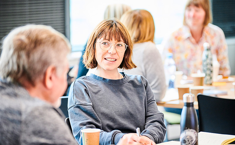 Light-skinned woman wearing glasses speaks to a light-skinned man during a Newcastle University Business School Executive Education workshop. They are seated at a table with notebooks, drinks, and a water bottle.