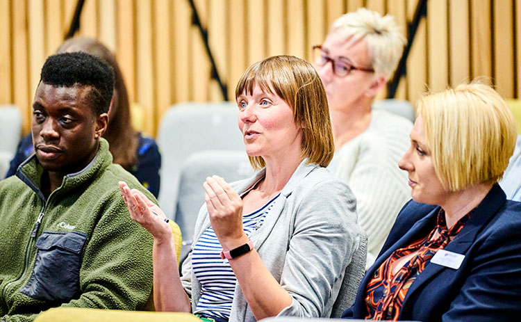A woman in a striped top and blazer speaks and gestures while seated in an auditorium. Other attendees, including a man in a green fleece and two women, listen.