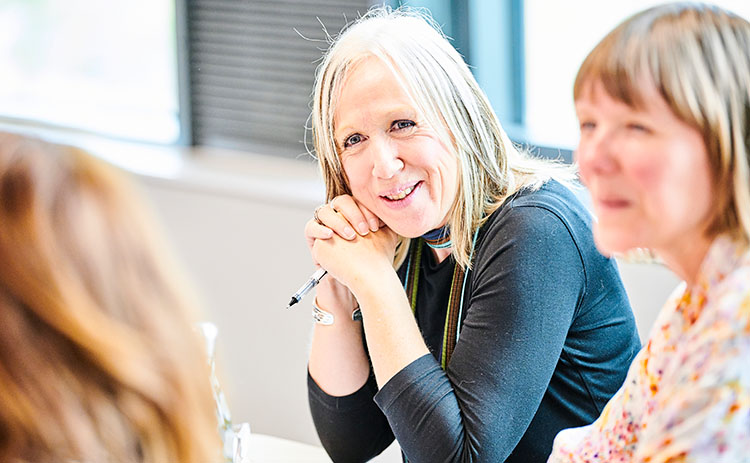 A light-skinned woman with long blonde hair smiling and holding a pen, engaged in a classroom discussion during an executive education session. Two other women, also light-skinned, sit beside her, partially visible. The background shows a bright, well-lit room.
