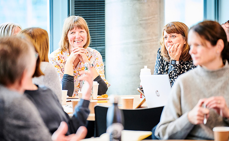A group of light-skinned women and men taking part in a Newcastle University Business School Executive Education workshop. They are seated around tables, talking and smiling, with drinks and notebooks in front of them.