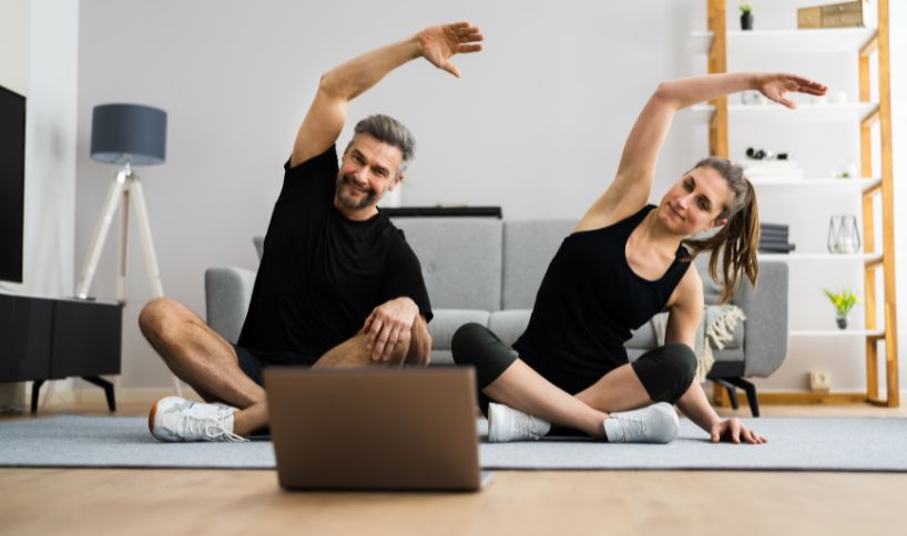 Man and woman in sportswear doing a seated side stretch while following an online workout on a laptop.