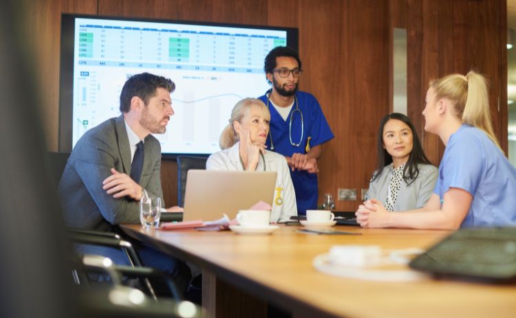 A mixed group of healthcare and business professionals meet around a table in a hospital boardroom, discussing data shown on a large screen.