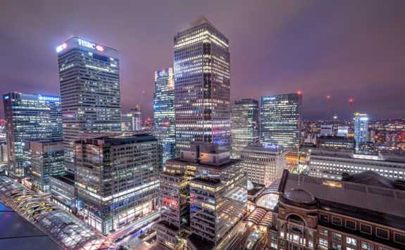 A night view of Canary Wharf in London, showcasing illuminated skyscrapers and banks against a dusky sky.