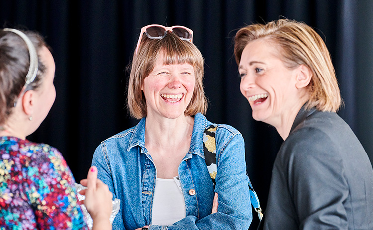 Three women laughing and chatting indoors. Jo Craggs, in a denim jacket with sunglasses on her head, is in the centre. Jaclyn Wright, with her back to the camera, wears a colourful top. A third woman in a dark blazer stands on the right.