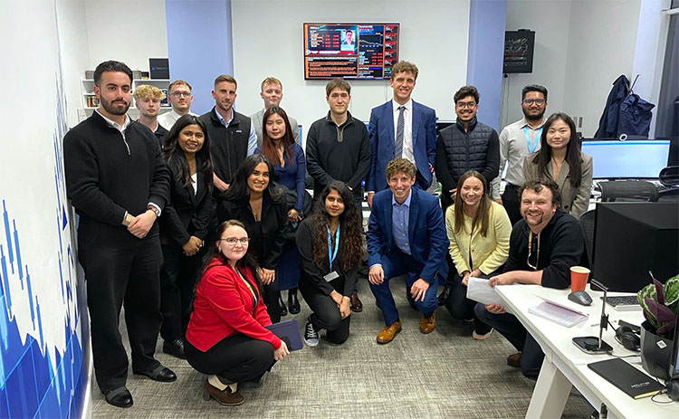Group photo of students and AmplifyME staff inside a financial training room. The group is dressed in business and casual attire, with financial data displayed on screens in the background