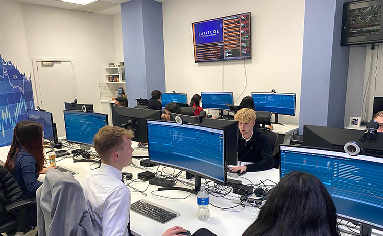 Students seated at curved monitors in a financial trading room at AmplifyME, participating in a market simulation. Blue data screens and market news displayed on monitors.