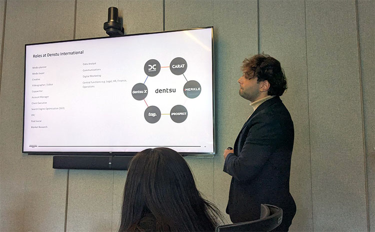 A Dentsu presenter in business attire delivers a presentation to students, with a slide showing company roles and logos displayed on a large screen.