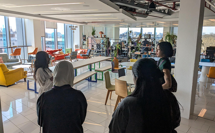 A group of students tours a modern, open-plan office with colourful seating areas, large windows, and city views. An employee leads the group through the workspace.