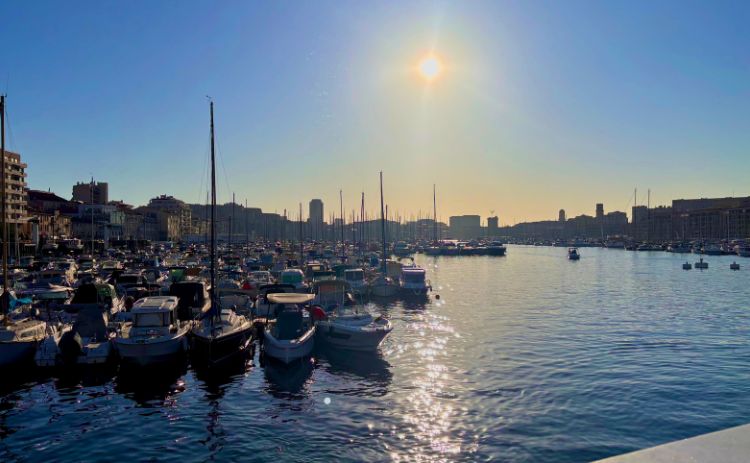 Scenic view of a marina filled with boats at sunset in Marseille, France, with the sun low in the sky and city buildings silhouetted in the background.