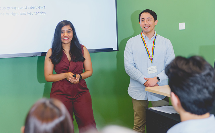 Two MBA students stand in front of a green wall and projected screen, presenting to an audience during a group project session at Newcastle University Business School.