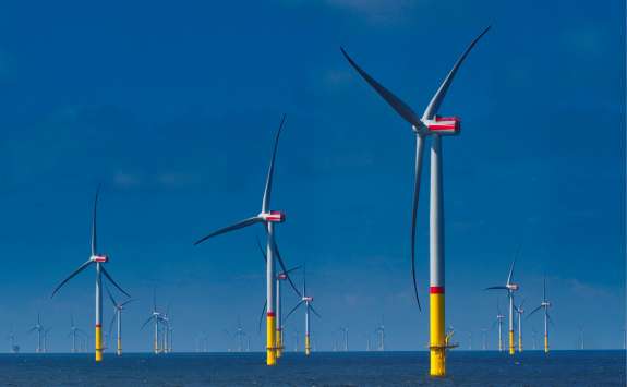 A series of large offshore wind turbines stand in the sea under a clear blue sky, with yellow and red markings on their bases