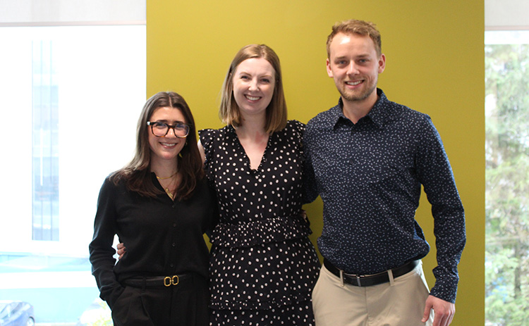 L-R: Francesca Principe, Pip Kyle, and Thomas Leaf stand smiling together indoors against a yellow wall.