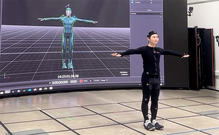A student in a motion capture suit stands in a T-pose in front of a large screen displaying a digital avatar mirroring their movements during a demonstration at the virtual production studio.