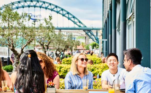 Business School students sat at a cafe table on the Newcastle Quayside with the Tyne Bridge in the background