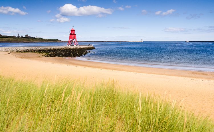 View of the River Tyne at South Shields with a red lighthouse on the pier, golden sandy beach, and grassy dunes under a blue sky with scattered clouds.