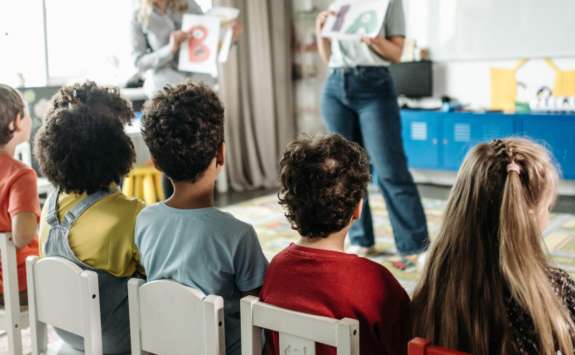 A group of schoolchildren sits in white chairs, facing two teachers holding up letters. The children are engaged and attentive, with their backs to the camera. The classroom is bright and decorated, creating a lively atmosphere.