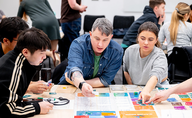 A group of students work together around a table, engaging with a colourful board game.