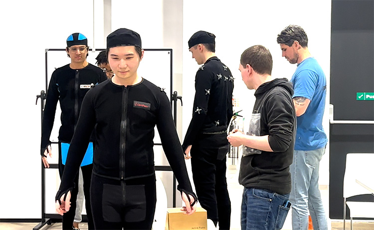 Students wearing black motion capture suits get ready for a virtual production session. They are assisted by technicians and stand in a brightly lit studio.