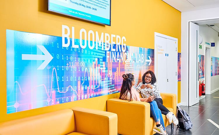 Two students sitting on a yellow sofa in front of a large colourful sign reading "Bloomberg Financial Lab" with an arrow pointing right.