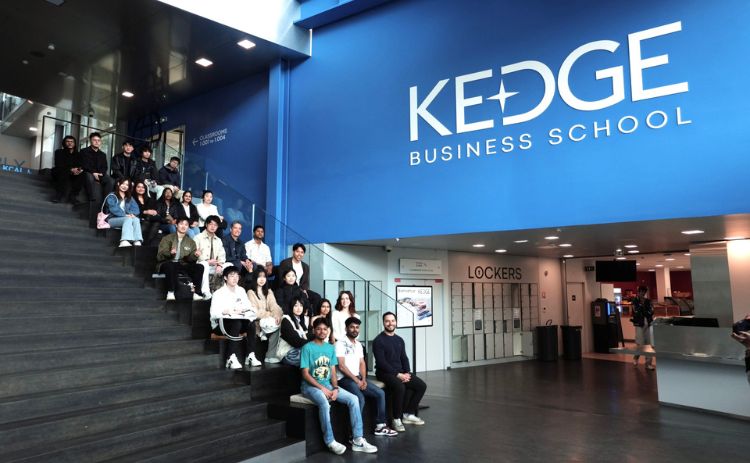 A group of students sits on wide indoor stairs beneath a large blue wall featuring the KEDGE Business School logo, inside a bright, modern building.
