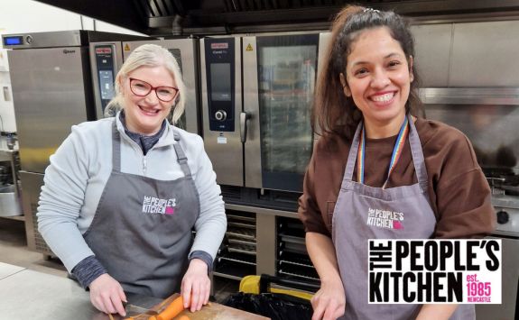 Sue Orr and Sonia Kapoor smiling in a commercial kitchen setting. They are chopping vegetables and wearing aprons with "The People’s Kitchen" logo.	