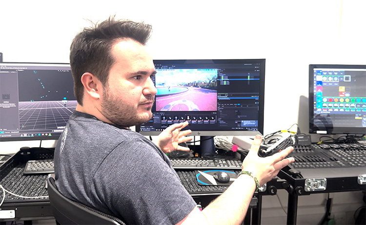A technician operates a virtual production control desk, gesturing while explaining the interface displayed on three monitors, including a simulation of a moving vehicle and digital environment settings.