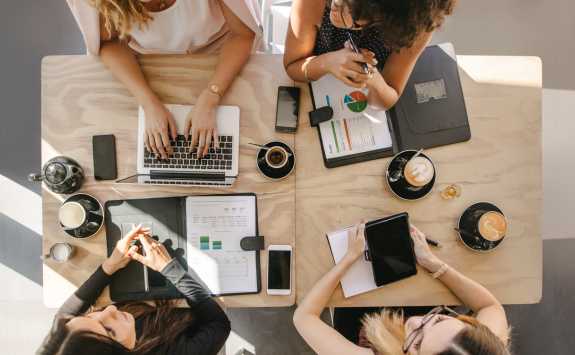 Four women entrepreneurs at a meeting table with laptops, tablets, and coffee, engaged in collaborative work, viewed from above.