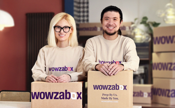 Wowzabox co-founders Georgina and Joshy Jin stood wearing company-branded sweatshirts and resting their arms on branded boxes