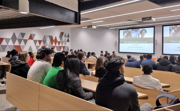 A photograph taken at the back of a large lecture theatre with an audience of mostly Black students. There is a screen at the front of the room which says