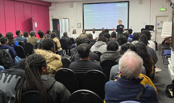 A room of majority Black students sit on chairs facing away from the camera. At the front of the room are five Black people who are on the network panel. There is a white woman standing in front of a projector screen next to the panellists.