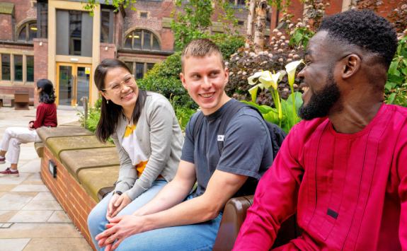 A group of three students sit together on a bench outside, laughing. One student is a black male, one student is a white male, and one student is an Asian female. There is a female sitting in the background facing away from the camera.